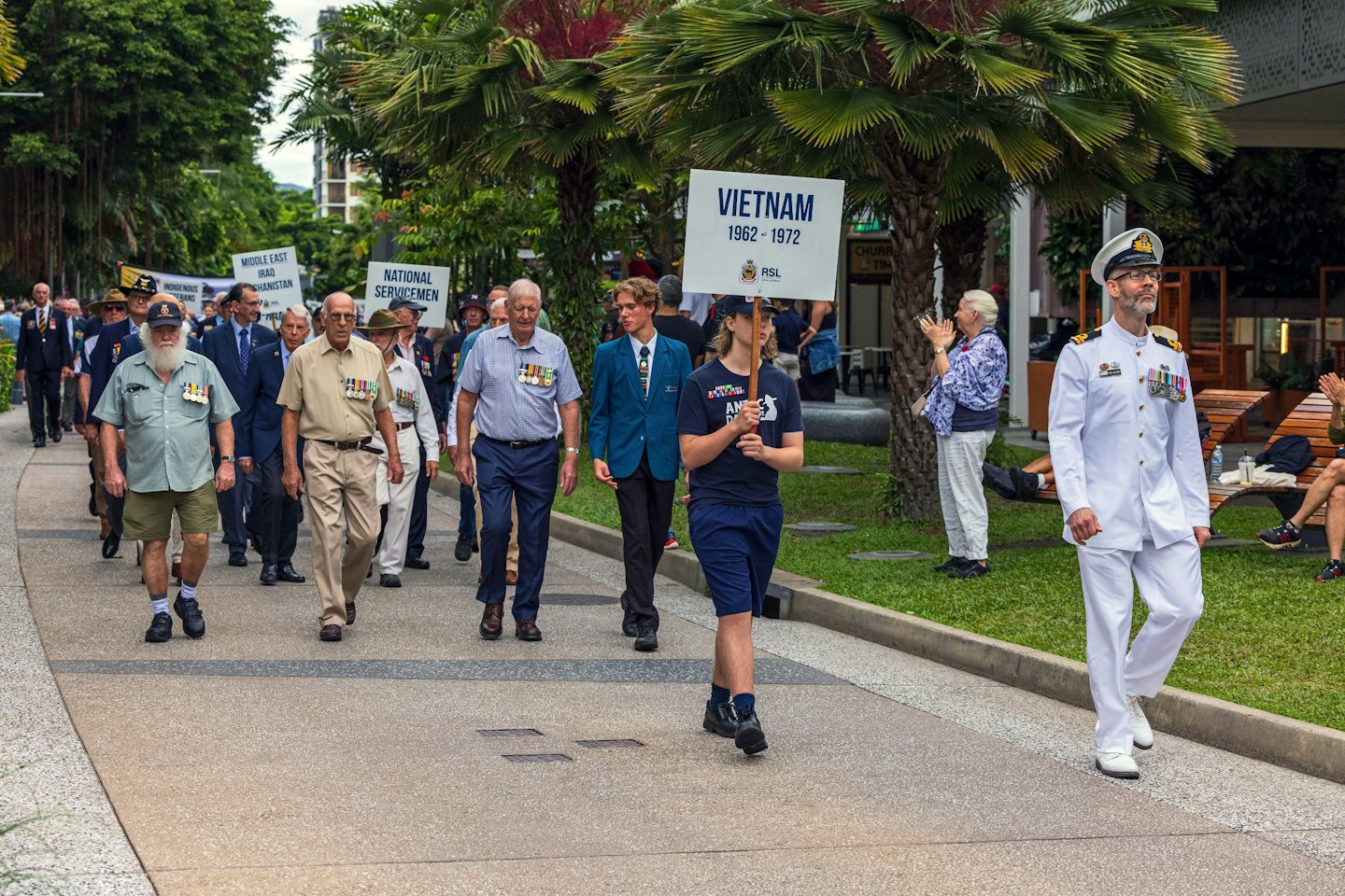 Military veterans march in a parade past palm trees.