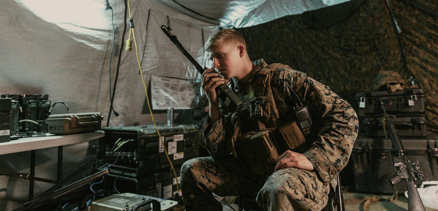 Man in military uniform speaks into radio in a field tent.